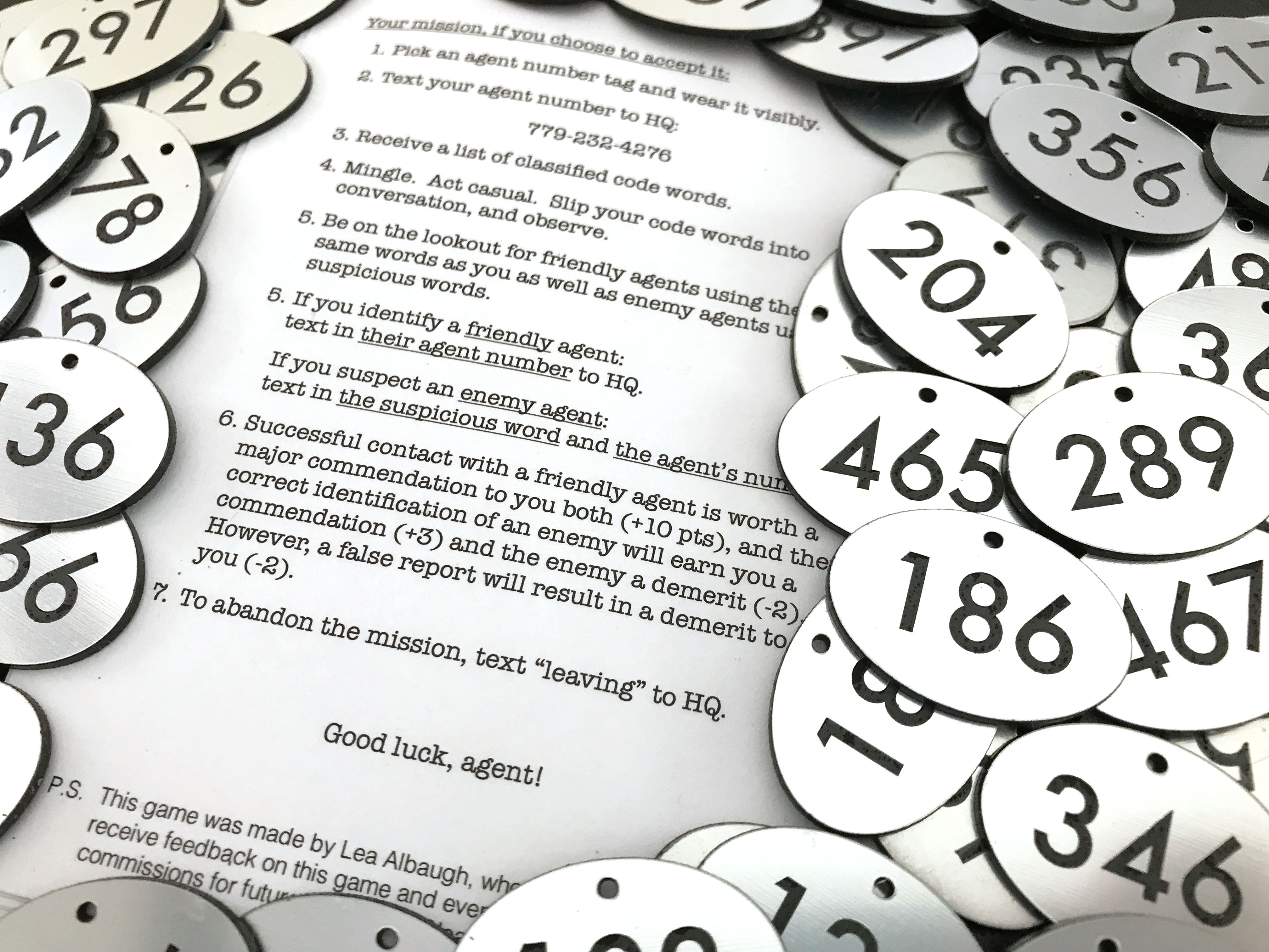 A game instruction sheet and laser-cut agent number badges.  The instruction sheet reads: "1. Pick an agent number tag and wear it visibly. 2. Text your agent number to HQ:
(number obscured) 3. Receive a list of classified code words. 4. Mingle. Act casual. Slip your code words into conversation, and observe. 5. Be on the lookout for friendly agents using the same words as you as well as enemy agents using suspicious words. 5. If you identify a friendly agent: text in their agent number to HQ. If you suspect an enemy agent: text in the suspicious word and the agent’s number. 6. Successful contact with a friendly agent is worth a major commendation to you both (+10 pts), and the correct identification of an enemy will earn you a commendation (+3) and the enemy a demerit (-2). However, a false report will result in a demerit to you (-2). 7. To abandon the mission, text “leaving” to HQ. Good luck, agent!"